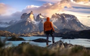 A hiker dressed in bright orange standing on a rocky hill overlooking the stunning Cuernos del Paine mountains in Torres del Paine, Chile, at sunrise. The vibrant colors of the sky and the turquoise waters below create a mesmerizing scene of Patagonia's natural beauty.